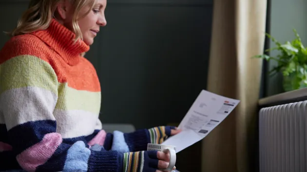 person trying to keep warm by a radiator looking at a utility bill 