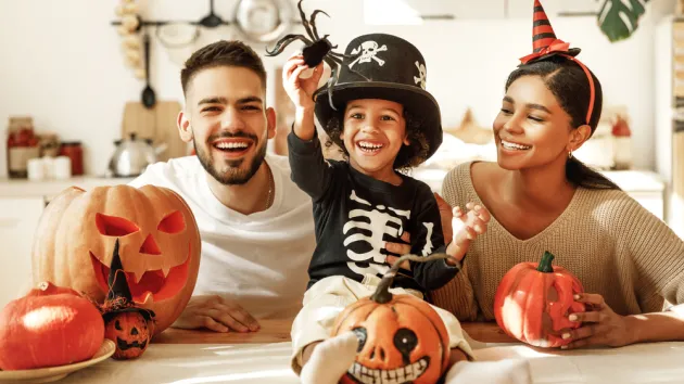  parents with son smiling while creating jack o lantern from pumpkin during Halloween celebration in kitchen at home