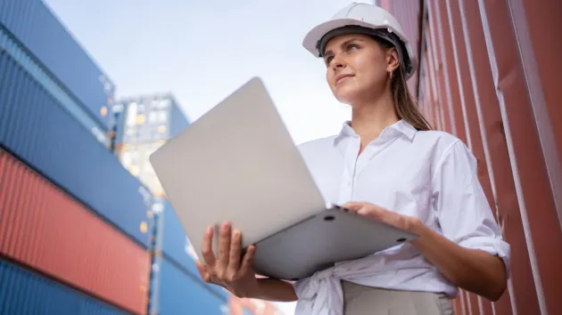 manager or engineer worker in casual suit standing in shipping container yard holding laptop