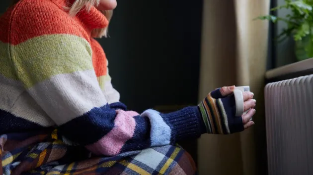 Woman In Gloves With Hot Drink Trying To Keep Warm By Radiator During Cost Of Living Energy Crisis