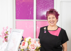 Wendy standing in front of a pink stained glass window in her black and pink therapies uniform