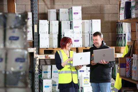 Man and woman in warehouse surrounded by product shelving holding laptops and looking 