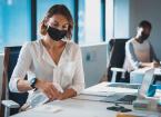 Two businesswomen wearing face masks sitting in office, one disinfecting her desk. Independent creative business at a modern office during coronavirus covid 19 pandemic.