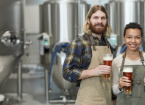 Waist up portrait of two smiling young workers holding beer glasses and looking at camera while standing in workshop at brewing factory