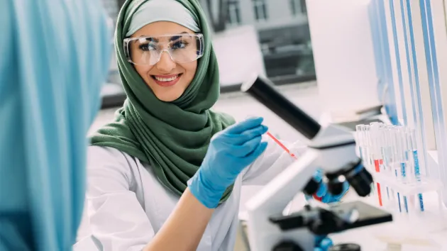smiling female Muslim scientist in goggles and hijab during experiment in chemical laboratory