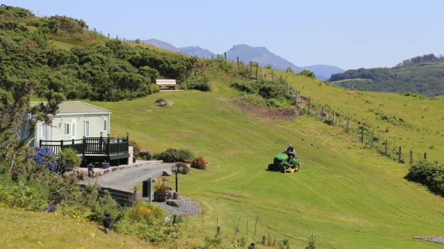 Criccieth, Gwynedd, Wales, a man using his ride on mower to cut the grass on a hillside around his cabin.