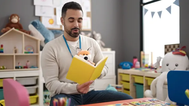 Young man preschool teacher reading story book sitting on table 