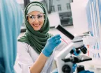 smiling female Muslim scientist in goggles and hijab during experiment in chemical laboratory