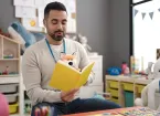 Young man preschool teacher reading story book sitting on table 
