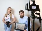 Stephen and Lyndsay sitting in front of studio back drop with camera in front of them and them holding a tablet displaying their website