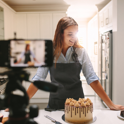 Woman in apron with cake in front of her and a smartphone on a tripod in front filming 