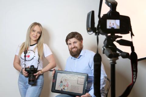 Stephen and Lyndsay sitting in studio in front of back drop showing their website on a tablet